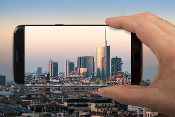 Canvas Print - Hand using smartphone taking photo of Milan at sunset, panoramic view with new skyscrapers of Porta Nuova district.