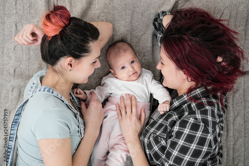 Two young women, a lesbian homosexual couple, are lying on a blanket with a child. Same-sex marriage, adoption.
