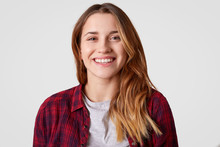 Close Up Shot Of Pretty Young Woman With Healthy Skin, Broad Smile, Shows White Teeth, Wears Checkered Shirt, Being In High Spirit, Looks Gladfully Directly At Camera, Hears Something Positive
