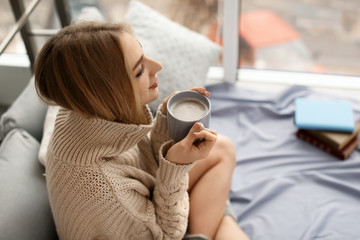 Sticker - Beautiful young woman in knitted sweater sitting with cup of coffee at home. Winter atmosphere