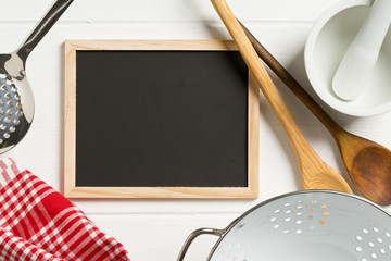 Wall Mural - Blank, empty, black chalkboard with cooking utensils and red checkered dish towel flat lay from above on white wooden table