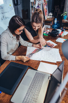 Young female graphic designers sitting at desk with laptop, color charts and some sketches discussing the new project choosing a concept