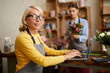 © Seventyfour - Side view portrait of mature female small business owner using laptop and looking at camera in flower shop, copy space