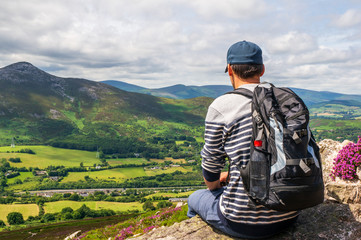 Tourist carrying a backpack, resting after a hike and enjoying the beautiful view of the green rolling Irish hills and mountain peaks. View from Little Sugar Loaf summit, Wicklow Mountains, Ireland.