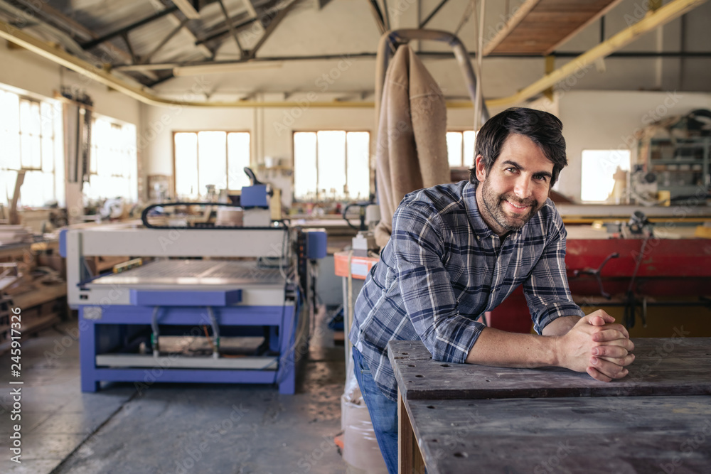 Woodworker smiling while leaning on a bench in his workshop - obrazy, fototapety, plakaty 