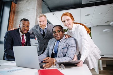 Close-knit business team: two African men with a red-haired girl and a gray-haired adult man sitting at a table in front of a laptop in a modern office