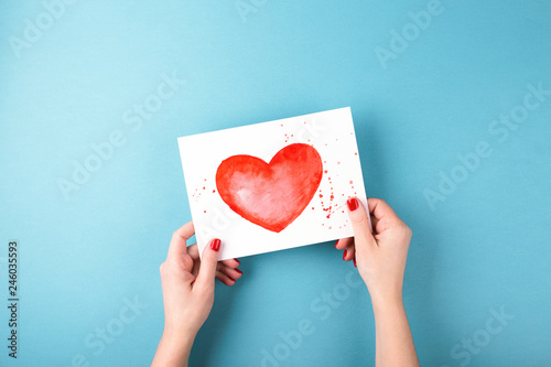 Overhead view on female hands holding card with a painted heart on blue backg...