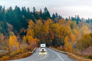 White big rig semi truck transporting goods on the winding autumn wet raining road at twilight