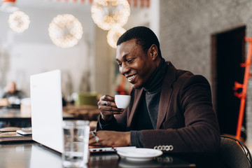 Portrait of happy african businessman using phone and drinking coffee while working on laptop in a restaurant.