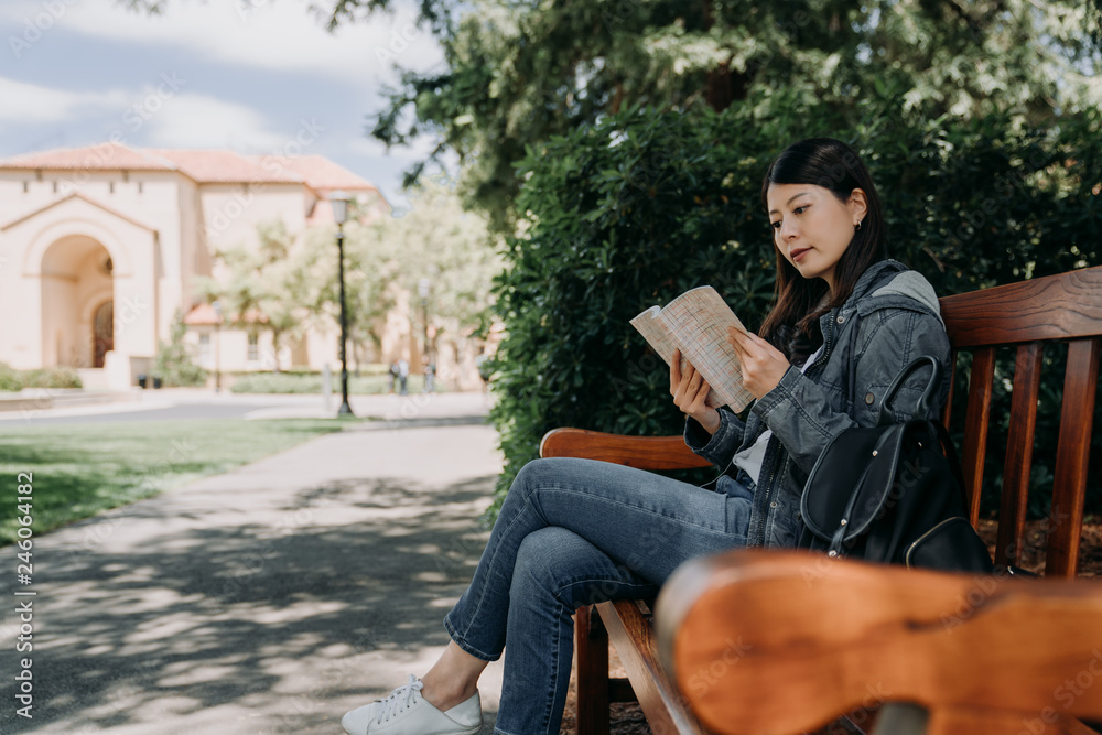 asian female college student sitting on wood bench reading novel book relaxing under tree shadows on sunny day outdoor. young girl studying abroad join short term summer tour program in usa stanford. - obrazy, fototapety, plakaty 
