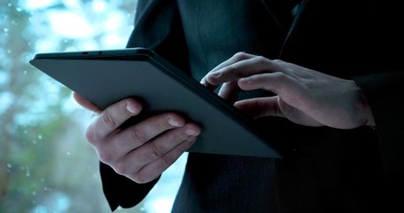 A man (guy) in a suit in a office building on the background of the street is holding a tablet. Concept from: Waiting, Meeting, Marketing, Making a meeting, Looking at a tablet, Writing a message.