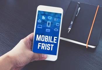 Poster - Hand holding mobile phone with mobile first word and feature icon over notebook on black wood table background,Digital marketing concept ,office desk..