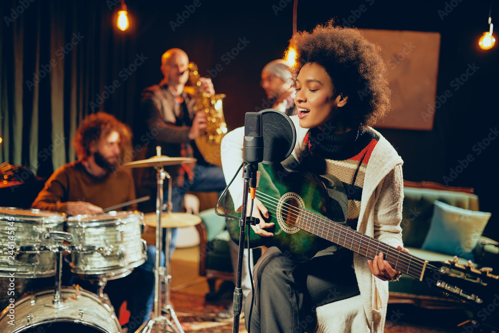 Mixed race woman singing and playing guitar while sitting on chair with legs crossed. In background drummer, saxophonist and bass guitarist. - obrazy, fototapety, plakaty 
