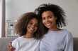 © fizkes - Portrait of happy African American mother and daughter hug sitting on couch at home, smiling black mom or nanny embrace teenage girl, spending time together, looking at camera posing for picture