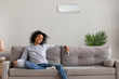 © fizkes - Calm African American woman sit on couch relaxing under air conditioner at home, peaceful black female hold remote controller resting on cozy sofa in apartment, happy girl enjoy fresh air