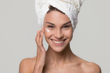 Smiling young woman apply moisturizing cosmetic cream on face look at camera with towel on head, happy girl care of healthy clean skin use spa treatment lotion isolated on studio background, portrait