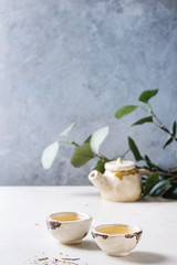 Hot green tea in two traditional chinese clay ceramic cup and teapot standing on white marble table. With green branch at background.