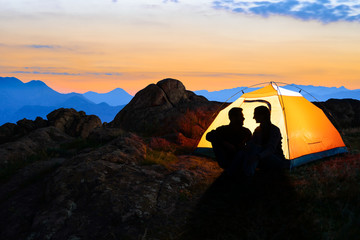 Wall Mural - young couple sitting near illuminated tent and looking at each other at beautirul evening in the mou