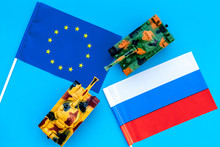 War, Confrontation Concept. European Union, Russia. Tanks Toy Near European And Russian Flag On Blue Background Top View