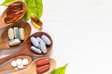 Variety of vitamin pills in wooden spoon on white background with green leaf, supplemental and healthcare product, flat lay surface