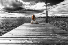 A Beautiful Girl Is Sitting Alone On The River Pier Against The Backdrop Of Thunder Clouds And Looks Into The Distance. View From The Back. The Wind Develops Her Curls Of Red Hair. 