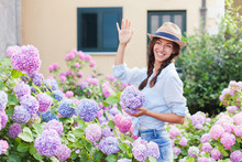 Girl Is In Bushes Of Hydrangea Flowers In Blooming Garden. Young Woman Greets Neighbors Or Welcomes Guests And Visitors. Concept Of Countryside Life Style, Gardening, Hospitality.