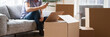 © fizkes - Close up carton boxes with belongings man sitting on sofa hold phone on background, wait call to delivery service moving at new house concept, banner for website header design with copy space for text
