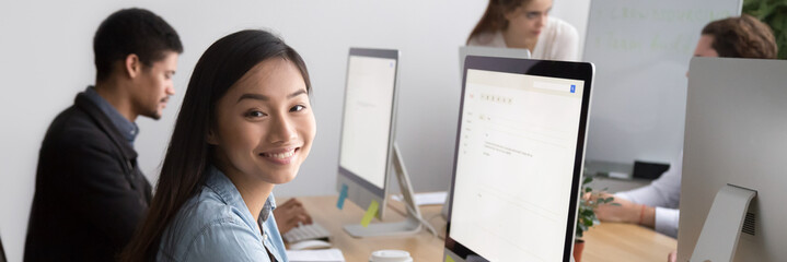 Smiling asian female worker looking at camera sitting at workplace working on pc with white mock up screen, diverse colleagues on background, banner for website header design with copy space for text