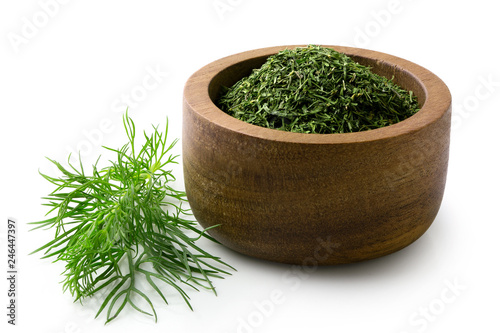 Fotografía Dried chopped dill in a dark wood bowl next to fresh dill leaves isolated on white
