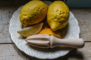 There is a white plate with two lemons and two orange slices on a wooden board table, next to a wooden stamp for squeezing juice.