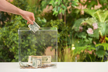 Man Putting Money Into Donation Box On Table Against Blurred Background, Closeup. Space For Text
