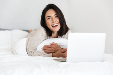 Wall Mural - Photo of adult woman 30s using laptop, while lying in bed with white linen in bright room