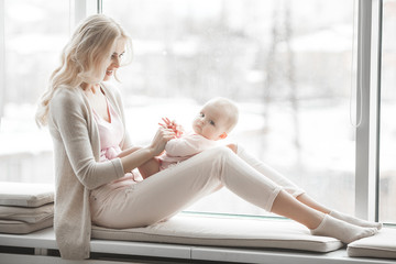 Wall Mural - Young mother taking care of her little baby girl. Beautiful mom and her daughter indoors in the bedroom. Loving family. Attractive mum holding her child.