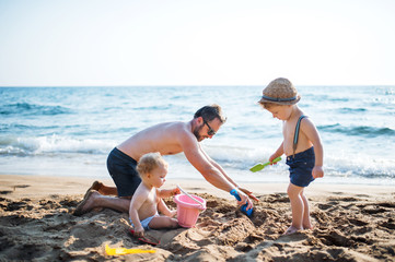 Wall Mural - A father with two toddler children playing with sand on beach on summer holiday.