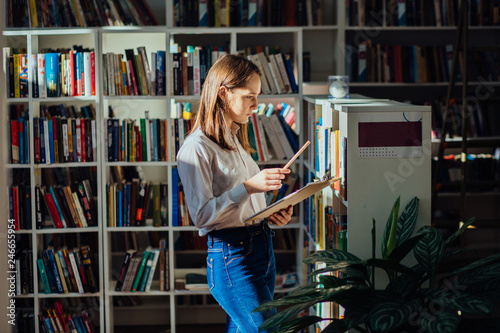 Student in library - smiling young female teenager search a book stand at bookshelf n college library Poster Mural XXL