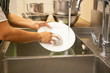 Hands washing dirty dishes with running water in kitchen sink.