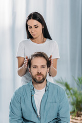 Wall Mural - bearded man looking at camera while receiving reiki treatment from young female healer