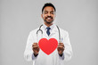medicine, cardiology and healthcare concept - smiling indian male doctor or cardiologist in white coat with red heart shape and stethoscope over grey background