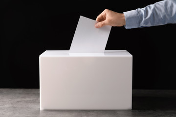 Man putting his vote into ballot box on table against black background, closeup