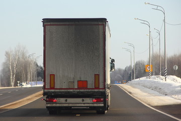 Logistics, international freight by road -  white european truck with semi trailer drive next to the two-lane asphalted suburban road in the winter day, rear view