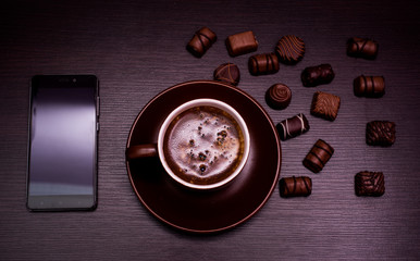 a cup of coffee, chocolate candy and a phone on a dark background.