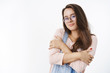 © Cookie Studio - Studio shot of tender silly young brunette in glasses and overalls hugging herself with dreamy pretty smile as if feeling chilly, wanting cuddles in warm hands standing against gray background
