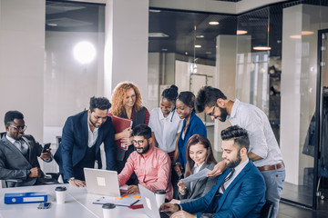 Wall Mural - Creative multiracial business team discussing project, standing around workplace, while two co-workers with portable computers looking for needed information, sharing results to the workers