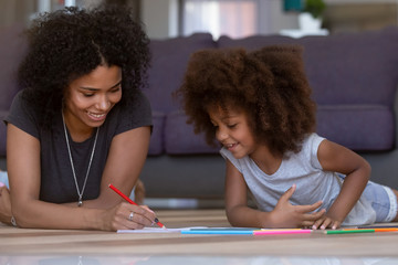 Wall Mural - Happy mixed race single mom and cute kid daughter draw with colored pencil on warm floor together, african mother baby sitter helping teaching child girl play have fun at home, creative family hobby