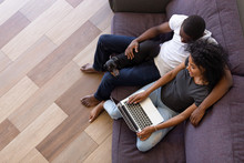 Happy Young African American Couple Enjoying Time At Home Using Computer Sitting On Sofa With Pet, Black Man And Woman Having Fun Online Shopping Or Surfing Web Relaxing On Couch With Dog, Top View
