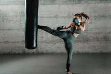 Caucasian Woman In Sportswear And With Boxing Gloves Kicking Bag In The Gym. Full Length. Wall In Background.