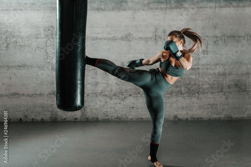 Fotomural Caucasian woman in sportswear and with boxing gloves kicking bag in the gym