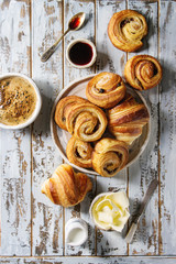 Variety of homemade puff pastry buns cinnamon rolls and croissant served with coffee cup, jam, butter as breakfast over white plank wooden background. Flat lay, space