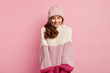 © wayhome.studio  - Horizontal shot of good looking woman with curly hair, looks with shy expression, wears warm hat and oversized knitted sweater, feels joyful, enjoys winter time, isolated over pink background.