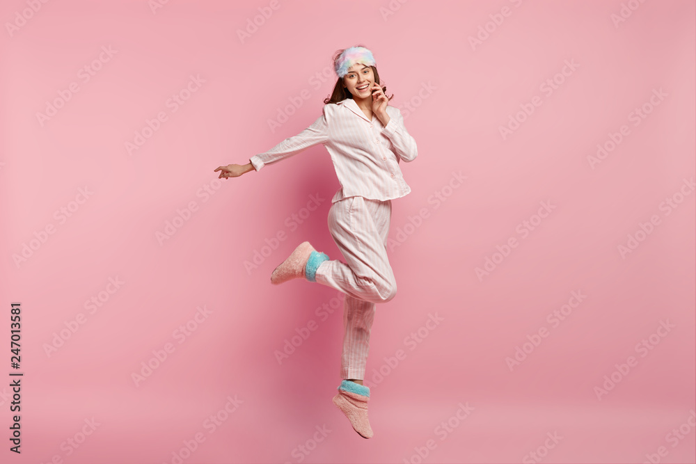 Indoor shot of overjoyed European woman raises leg, spreads hand, wears casual pyjamas and eyemask, smiles happily, isolated over pink background, enjoys bed time and good rest during weekend - obrazy, fototapety, plakaty 